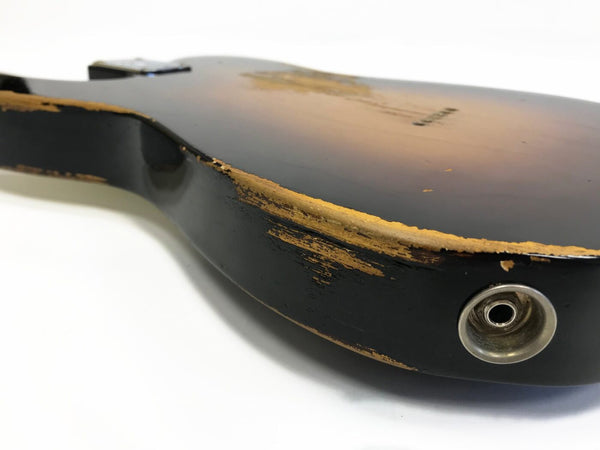Close-up of vintage electric guitar body with worn black and sunburst finish showing natural wood exposed and metal jack input socket on white background.