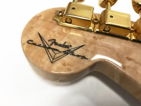 Close-up of Fender Custom Shop guitar headstock with natural wood finish and gold tuning pegs showing detailed logo and wood grain texture
