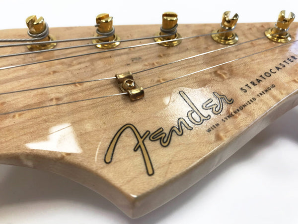 Close-up of Fender Stratocaster guitar headstock with gold tuning pegs and natural wood finish showing Fender logo and synchronized tremolo text