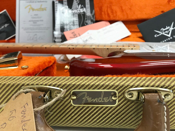 Fender custom guitar case with leather handles and metal nameplate, vintage tweed texture and certificate of authenticity in background, guitar neck and red guitar body visible inside orange plush lining case