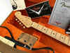 Close-up of Fender Stratocaster guitar headstock with gold tuning pegs in orange plush-lined guitar case, including Fender Custom Shop card, certificate of authenticity, and guitar strap.
