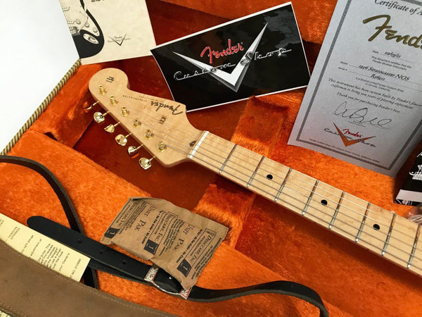 Close-up of Fender Stratocaster guitar headstock with gold tuning pegs in orange plush-lined guitar case, including Fender Custom Shop card, certificate of authenticity, and guitar strap.