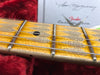 Close-up of maple guitar neck with black dot inlay and metal frets on six strings, resting on red plush case lining and blurred Fender certificate in background