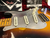 Close-up of vintage Fender Stratocaster electric guitar body with sunburst finish, white single-coil pickups, volume and tone knobs, and worn maple neck fretboard, accompanied by Fender owner's manual and certificate inside red plush case