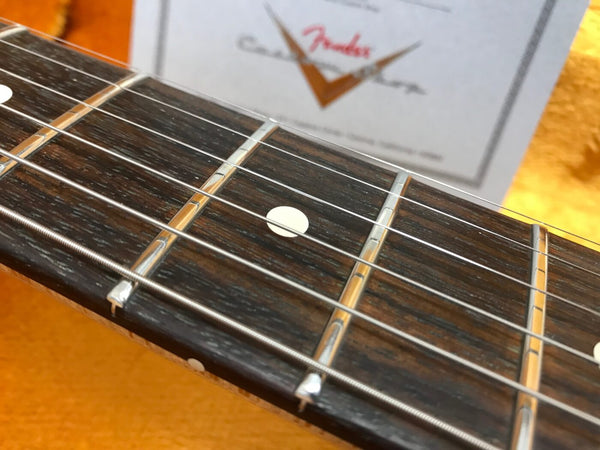 Close-up of Fender guitar rosewood fretboard with metal frets and white dot inlays against an orange background