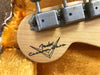 Close-up of Fender Custom Shop logo on natural wood guitar headstock with vintage tuning pegs and orange plush guitar case lining