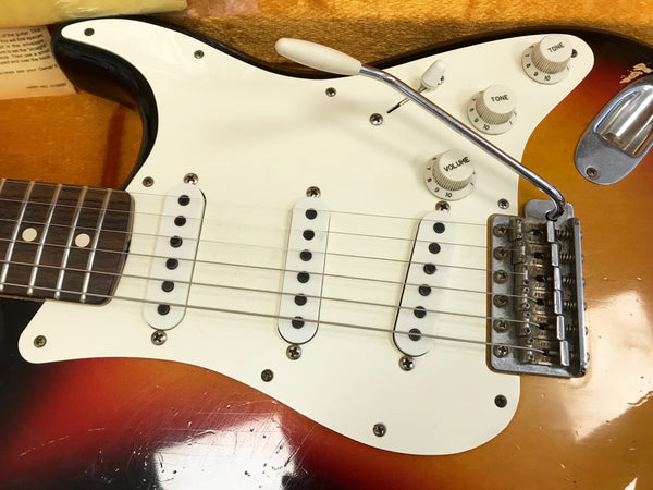 Close-up of vintage sunburst electric guitar body featuring white pickguard, three single-coil pickups, volume and tone control knobs, tremolo arm, and rosewood fretboard.