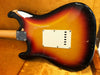 Vintage sunburst electric guitar body with worn finish and white backplate, displayed in an orange velvet-lined guitar case.