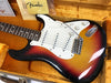 Vintage Fender Stratocaster electric guitar in sunburst finish with white pickguard, three single-coil pickups, and tremolo bridge in orange plush-lined hardshell case, including Fender cloth and documentation.