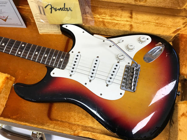Vintage Fender Stratocaster electric guitar in sunburst finish with white pickguard, three single-coil pickups, and tremolo bridge in orange plush-lined hardshell case, including Fender cloth and documentation.