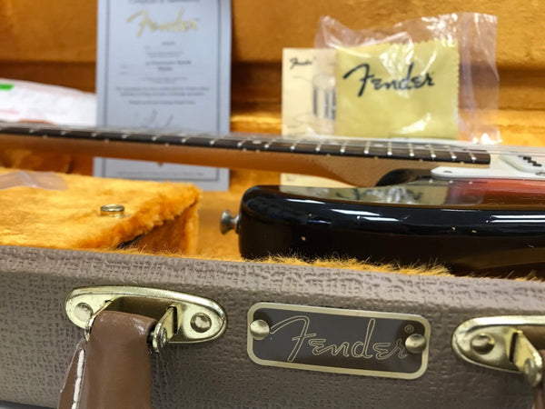 Close-up of Fender guitar case with brown leather handle and metal hardware, showing part of a black electric guitar inside with yellow plush lining and Fender branded certificate and accessories in the background.