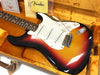 Vintage Fender Stratocaster electric guitar in sunburst finish with white pickguard and tremolo arm displayed in orange plush-lined guitar case with Fender cleaning cloth and certificate