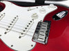 Close-up of red electric guitar body showing white pickguard, three single-coil pickups, volume and tone control knobs, and chrome bridge with strings, placed in guitar case