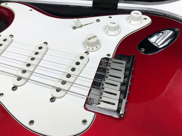 Close-up of red electric guitar body showing white pickguard, three single-coil pickups, volume and tone control knobs, and chrome bridge with strings, placed in guitar case