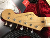 Close-up of Fender Stratocaster guitar headstock with tuning pegs, original contour body logo, and guitar cable on red plush background