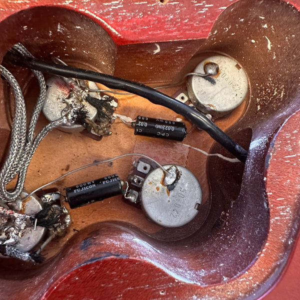 Close-up of vintage electric guitar control cavity showing aged potentiometers, capacitors, and wiring on wooden body with red finish.