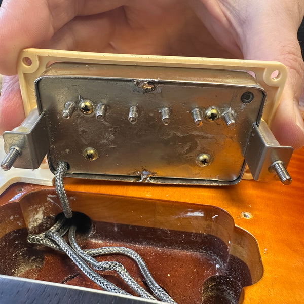 Close-up of vintage electric guitar pickup being held above guitar body cavity, showing metal backplate, mounting screws, and braided wiring in sunburst finish guitar
