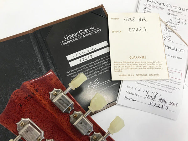 Gibson Custom Certificate of Authenticity for LP Standard guitar with serial number 87283 and guarantee card next to vintage guitar headstock with tuning pegs and serial number stamp visible, pre-pack checklist in background.