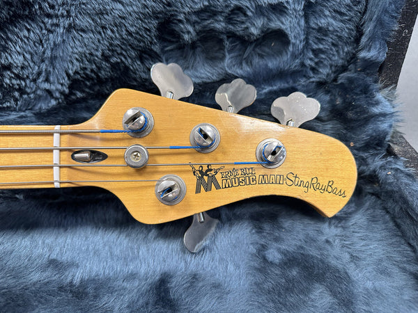 Close-up of Ernie Ball Music Man StingRay Bass guitar headstock with tuning pegs and strings in a plush blue guitar case interior.