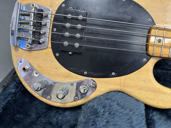 Close-up of vintage natural finish electric bass guitar body showing chrome control knobs, black pickguard, pickup, and strings resting on blue furry guitar case lining