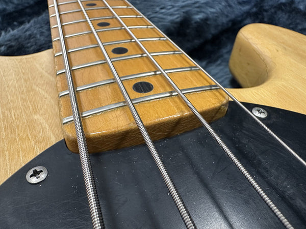 Close-up image of electric bass guitar maple fretboard with metal frets and black dot inlays, natural wood body, and black pickguard with screws