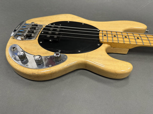 Natural wood finish electric bass guitar body with chrome control knobs and black pickguard on gray background