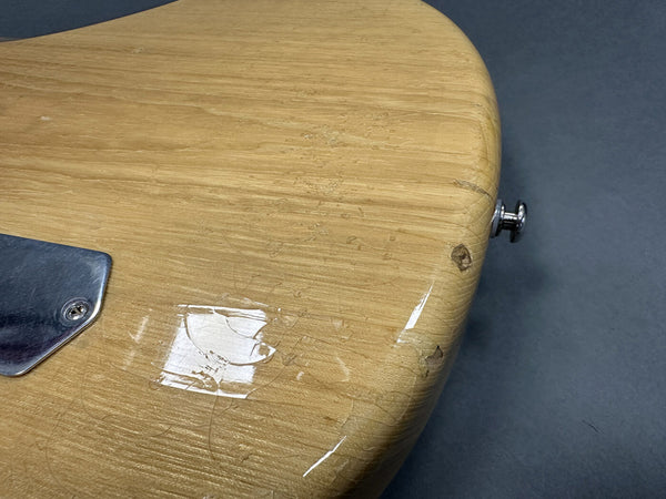 Close-up of worn light wood guitar body with visible scratches and chipped finish near strap button and metal plate on dark background