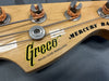 Close-up of Greco Mercury Bass guitar headstock showing tuning pegs, strings, and logo with "Brazen Picker Professional" text on natural wood finish.