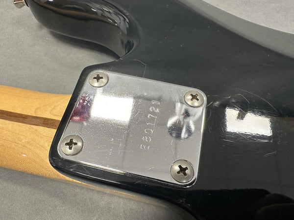Close-up of electric guitar neck plate with serial number E801721 on black guitar body and wooden neck, showing screw details and wood grain quality.