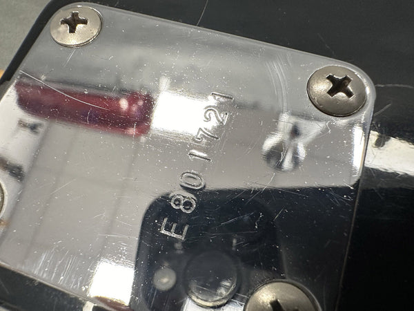 Close-up of a metal neck plate on a black electric guitar with serial number E801721 and four screws, showing some surface scratches and reflections