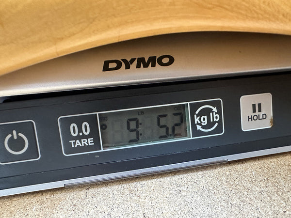 Close-up of DYMO digital scale display showing guitar weight of 9.52 pounds with tare, unit conversion, and hold buttons, scale placed on wooden surface