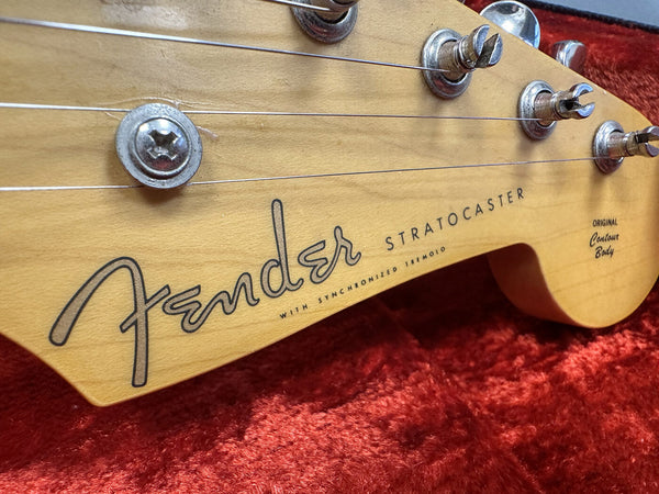 Close-up of Fender Stratocaster guitar headstock with tuning pegs and strings on red plush background, showing Fender logo and original contour body text.