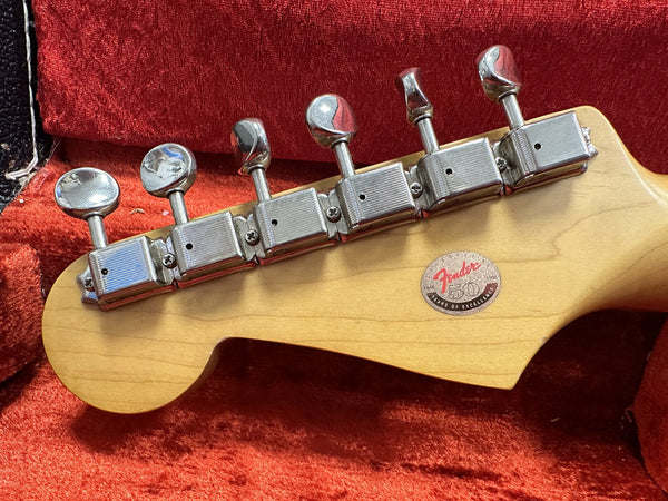 Close-up of Fender guitar headstock with vintage tuning pegs and 50th anniversary badge, maple wood finish, resting on red velvet guitar case lining