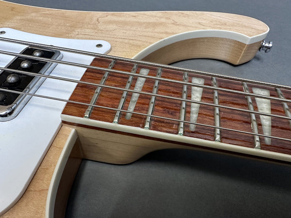 Close-up of electric bass guitar neck with natural wood finish, pearl trapezoid fretboard inlays, and chrome pickups on dark gray background