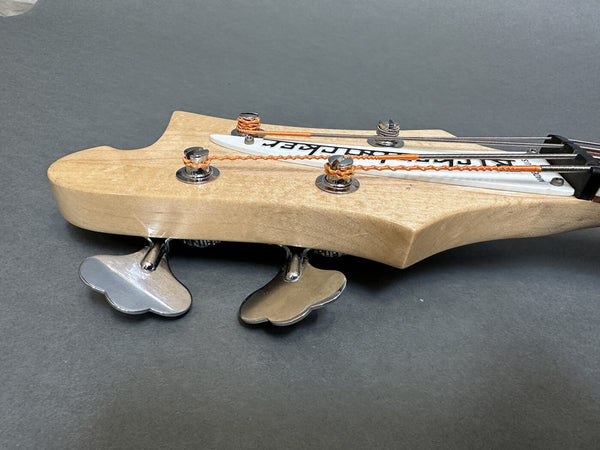 Close-up of maple bass guitar headstock with silver tuning pegs and orange-wrapped strings against gray background