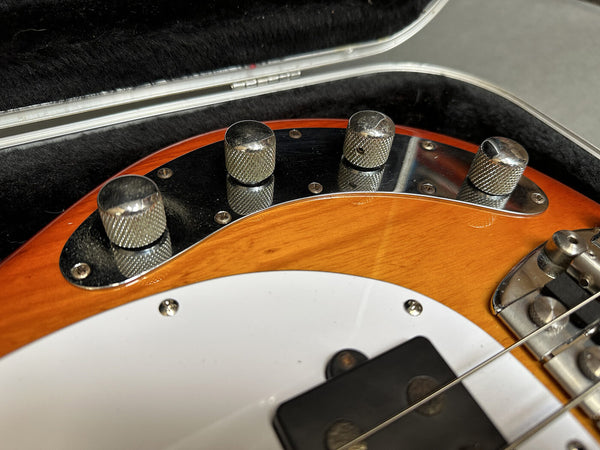 Close-up of maple body electric bass guitar with chrome control knobs, white pickguard, and black pickups in black plush case