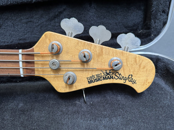 Close-up of Ernie Ball Music Man StingRay electric bass guitar headstock with tuning pegs and strings, natural wood finish.