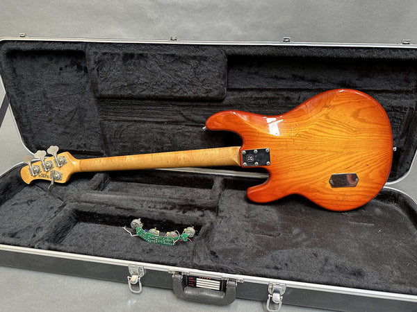 Vintage electric bass guitar with sunburst finish laying in black plush-lined hard case, showing back and neck with tuning pegs and removed electronic circuit board.
