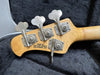 Close-up of bass guitar headstock with four tuning pegs and "Made in San Luis Obispo California USA" text on natural wood finish, resting on black guitar case lining.