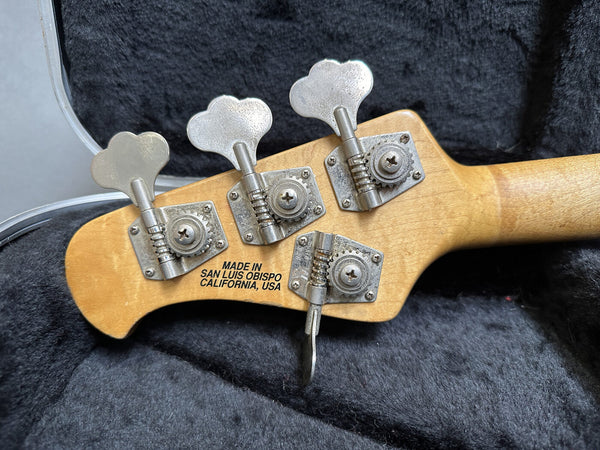 Close-up of bass guitar headstock with four tuning pegs and "Made in San Luis Obispo California USA" text on natural wood finish, resting on black guitar case lining.