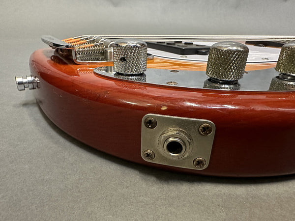 Close-up of vintage electric guitar body showing red wood finish, chrome input jack plate, control knobs, and bridge components on gray background.
