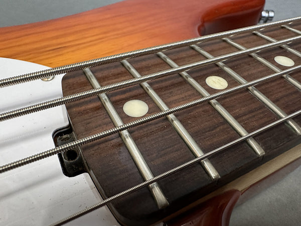 Close-up of bass guitar rosewood fingerboard with silver frets and round white inlays, showing thick metal bass strings on light wood neck and orange guitar body edge