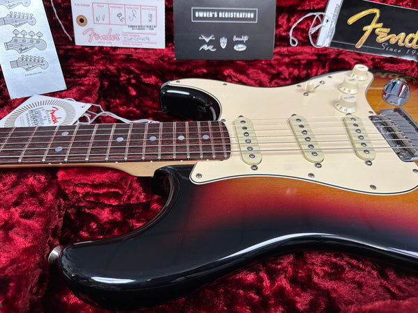 Close-up of Fender Stratocaster electric guitar with sunburst finish, rosewood fretboard, and three single-coil pickups, displayed in red velvet-lined case with Fender tags and owner's registration card