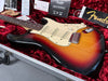 Fender Sunburst Stratocaster electric guitar in silver hard case with red plush interior, showing three single-coil pickups, white pickguard, tone and volume knobs, and Fender owner's manual and registration card.