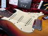 Fender Stratocaster electric guitar close-up showing sunburst finish, white pickguard, three single-coil pickups, volume and tone control knobs, and owner's registration card on red velvet background