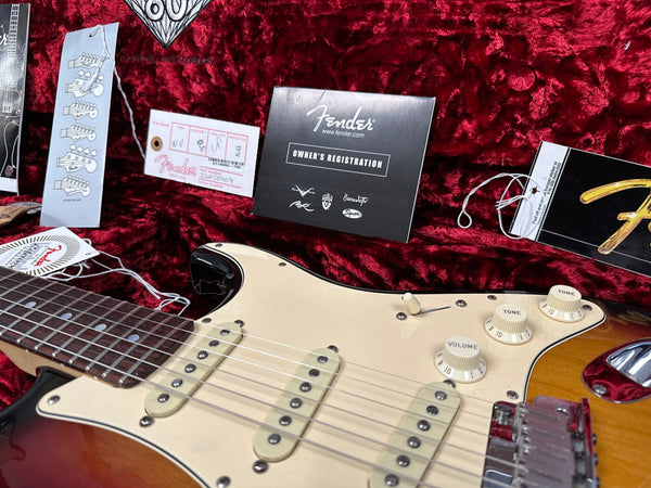 Fender Stratocaster electric guitar close-up with sunburst finish, white pickguard, tone and volume knobs, inside red plush case with Fender owner's registration card and guitar tags visible