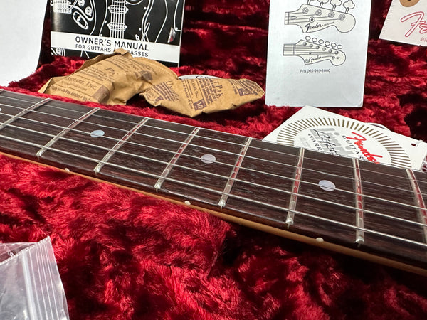 Close-up of a guitar fretboard with metal frets and dot inlays, surrounded by Fender owner's manual, warranty information, and silica gel packs on a red plush surface.