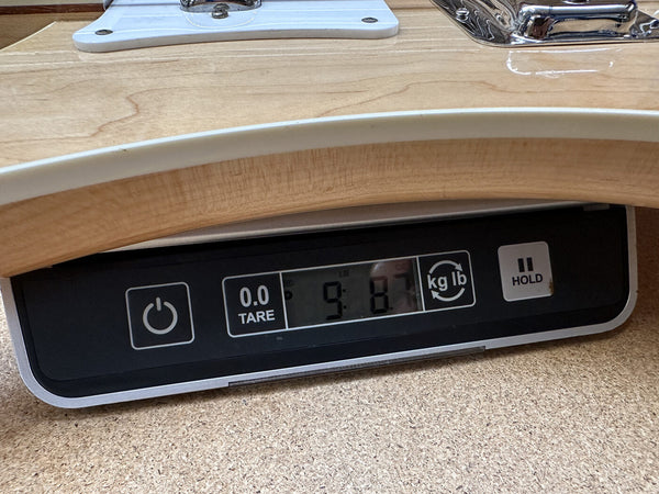 Digital scale showing weight of guitar at 9.87 pounds with close-up of natural wood guitar body edge and chrome hardware
