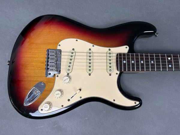 Electric guitar close-up showing sunburst finish, three single-coil pickups, volume and tone control knobs, and rosewood fretboard on gray background