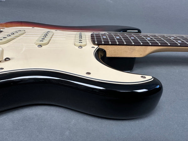 Close-up of black electric guitar body with cream pickguard and three single-coil pickups, rosewood fretboard, and maple neck on gray background